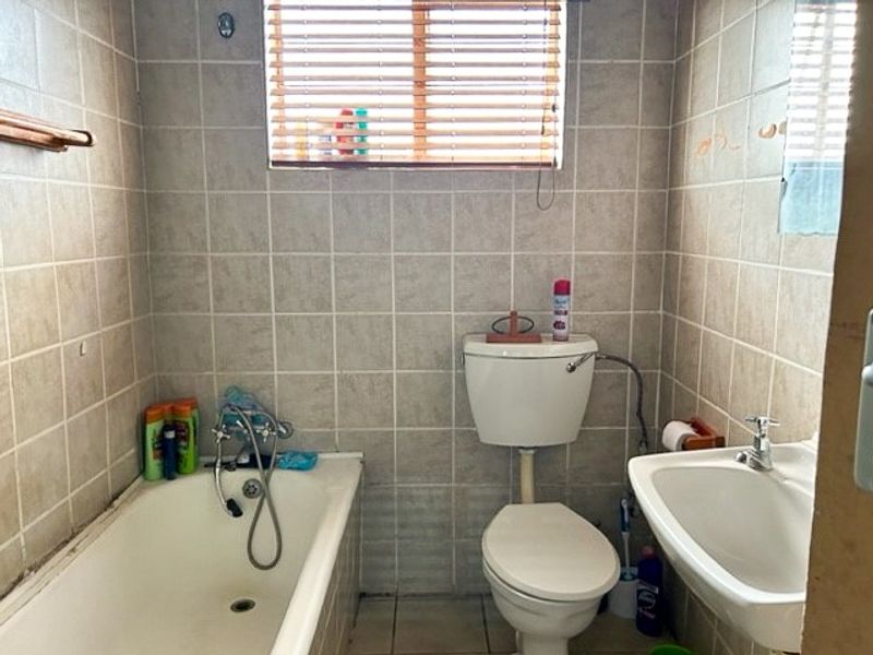 A functional bathroom featuring a standard bathtub, pedestal basin, and toilet, finished with neutral wall tiles and a window with fitted blinds.
