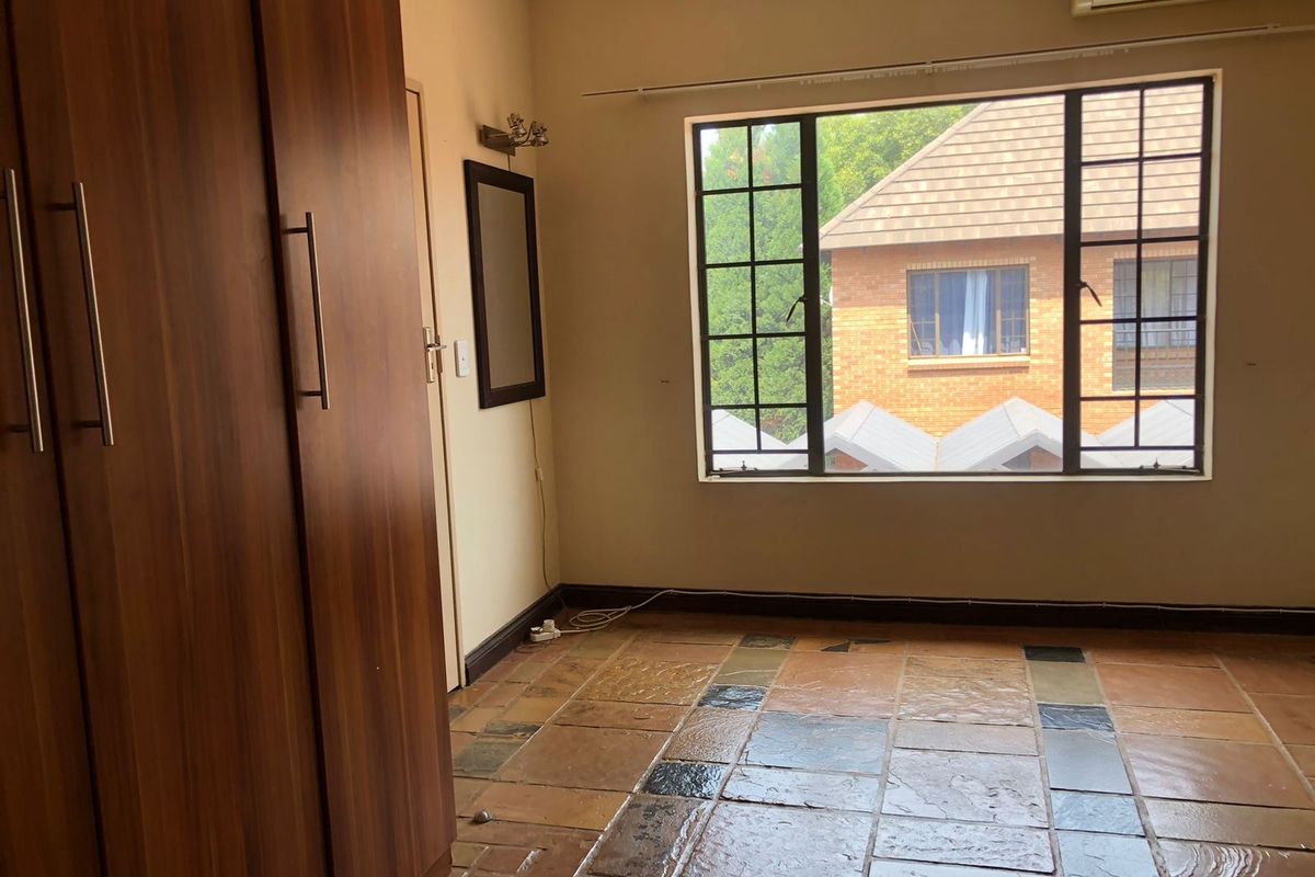 A bedroom featuring built-in wooden cupboards, tiled flooring, and a large window providing natural light and a view of the surrounding complex.
