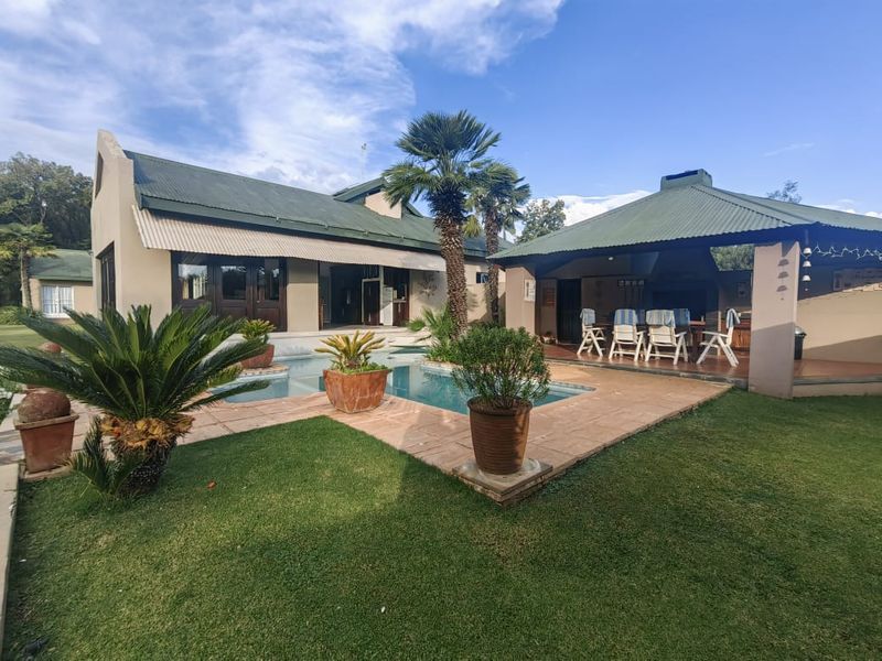 Outdoor entertainment area featuring a swimming pool, paved deck, and a covered patio with a built-in braai, set within a landscaped garden.