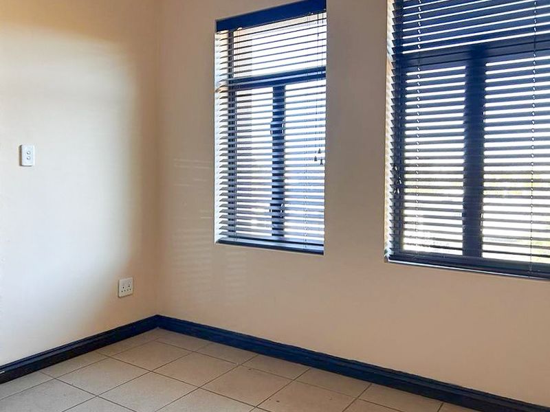 A bright, empty bedroom featuring neutral tiled flooring, large windows with fitted venetian blinds, and clean wall finishes.