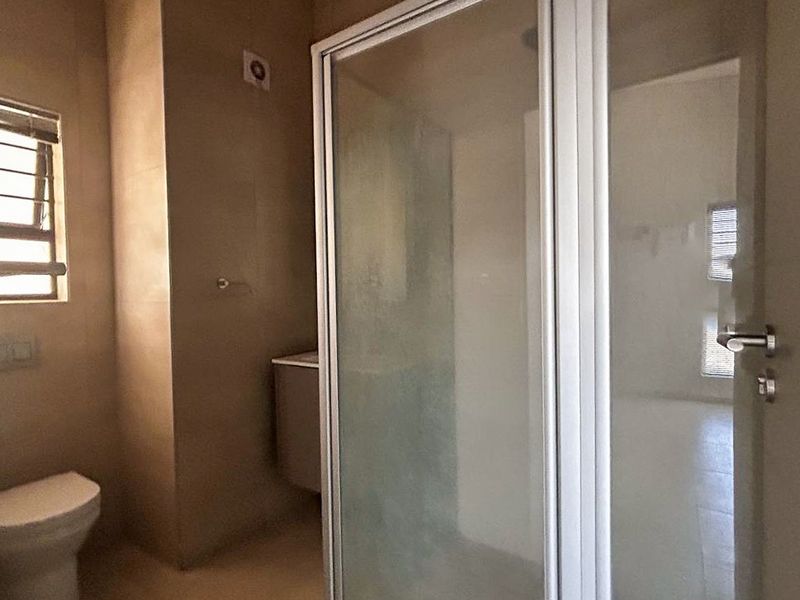 A functional bathroom featuring a glass-enclosed shower, a standard toilet, and neutral wall tiling with natural light from a side window.