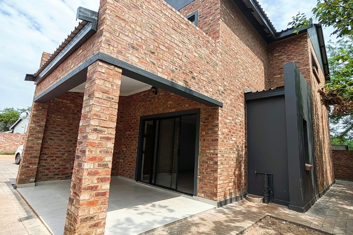 A modern double-story house featuring exposed face-brick exterior, a covered patio area with tiled flooring, and black-framed sliding glass doors.
