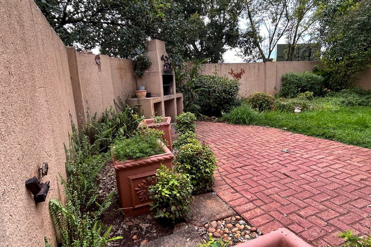 A private rear garden featuring a paved patio area, established landscaping, and a built-in brick braai fireplace against the boundary wall.