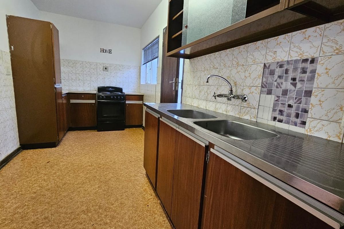 A dated kitchen featuring original wood-finish cabinetry, a double stainless steel sink, and tiled splashbacks, offering a functional space ready for modernization.