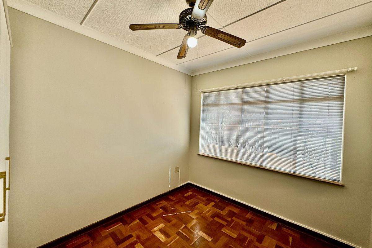 A bright, empty bedroom featuring classic parquet wood flooring, a ceiling fan, and large windows with fitted blinds.