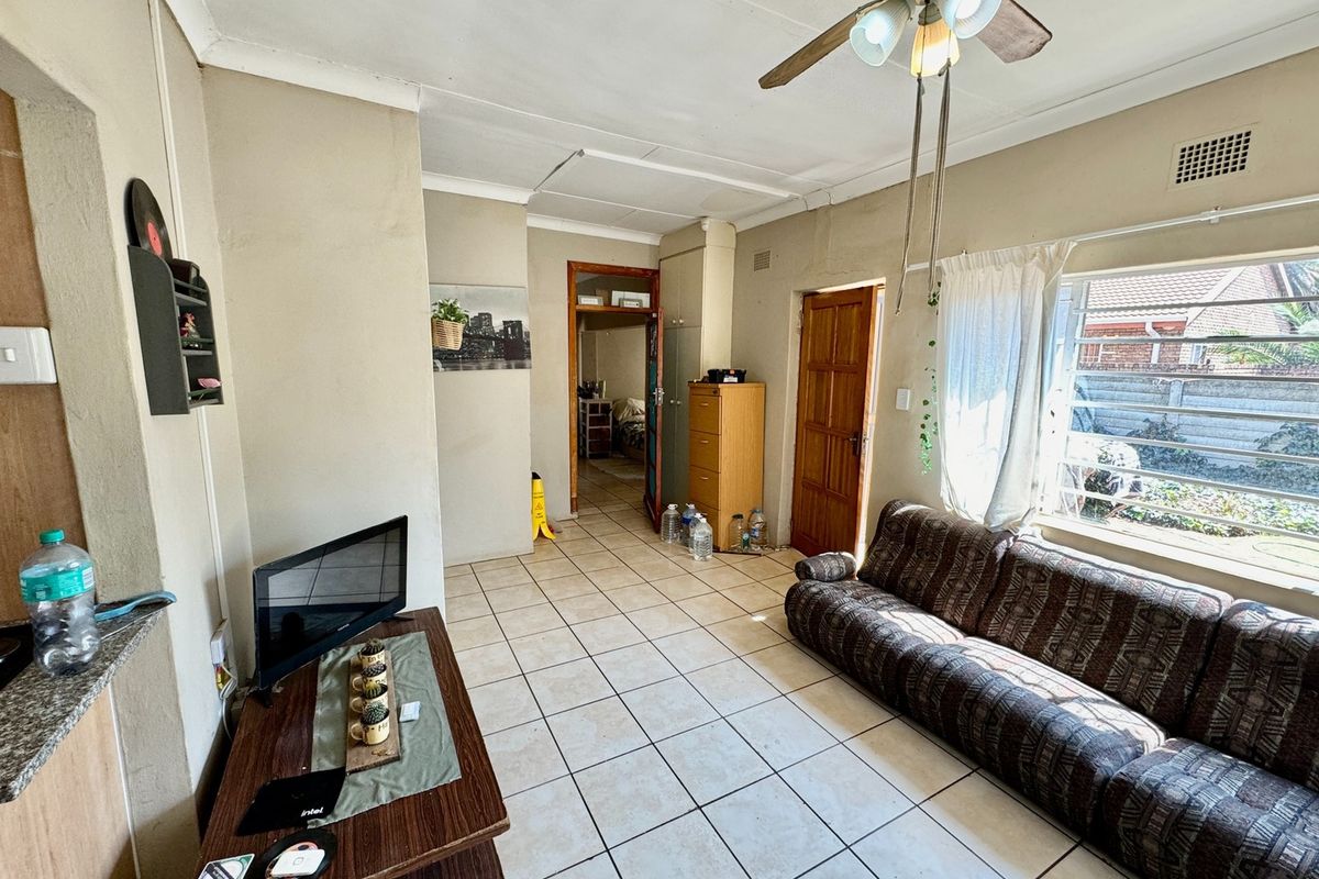 A living room featuring tiled flooring, a ceiling fan, and natural light from a large window, providing access to adjacent rooms.