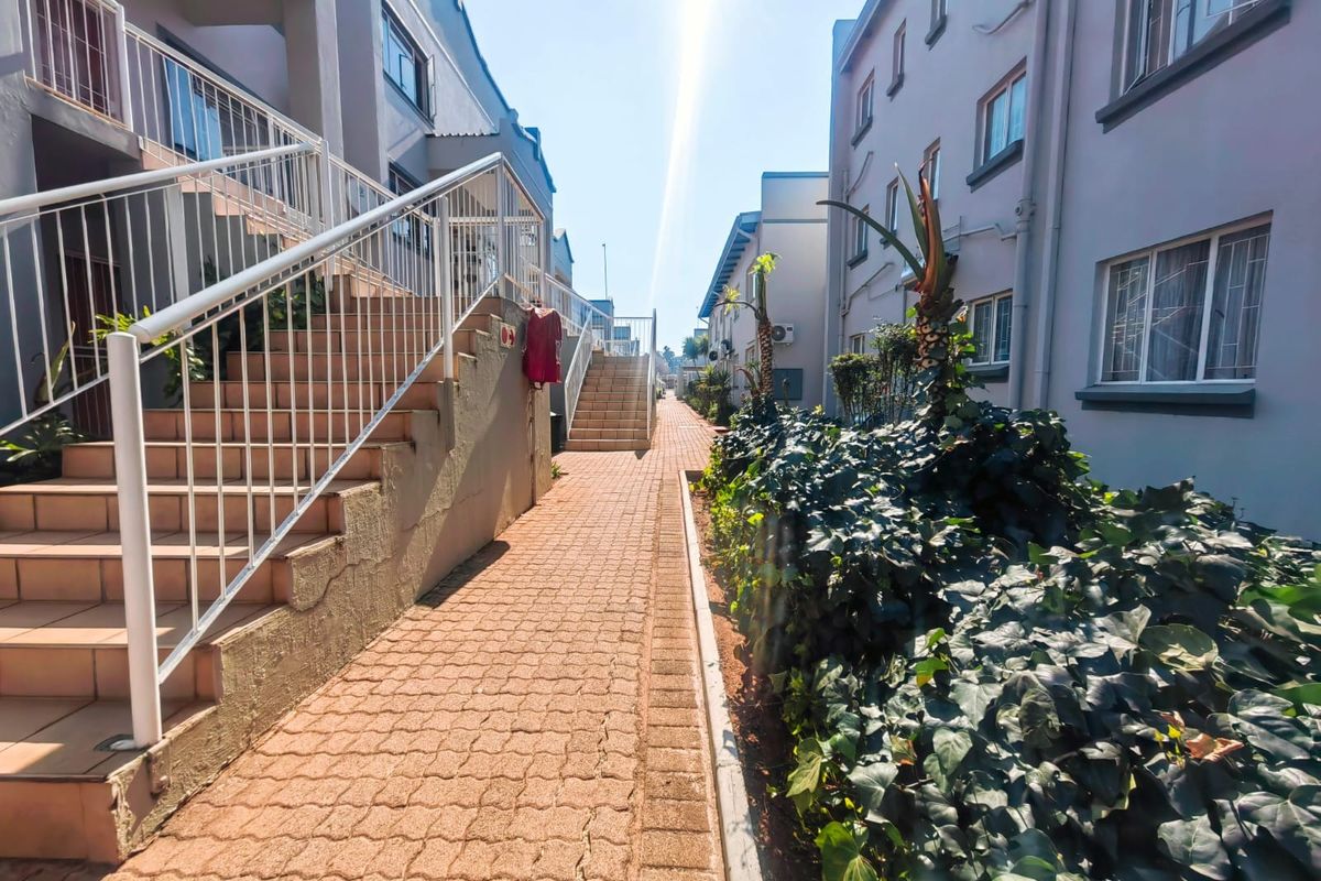 Exterior view of an apartment complex featuring paved walkways, external staircases with metal railings, and landscaped garden beds between the building blocks.