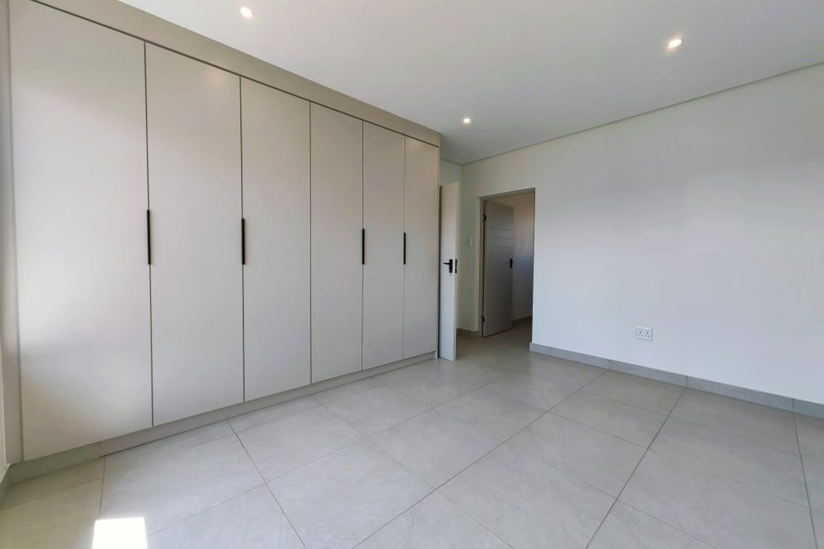 A bright, modern bedroom featuring neutral floor tiles, recessed ceiling lighting, and extensive built-in floor-to-ceiling cabinetry with sleek black handles.