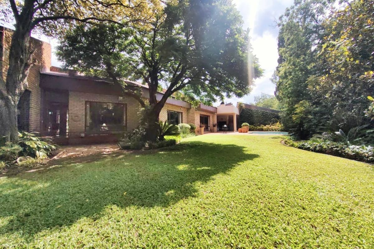 A spacious rear garden featuring a well-maintained lawn, mature trees, and a view of the property's rear facade and patio area.