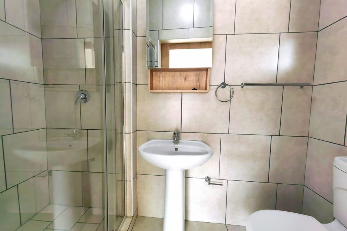 A functional bathroom featuring a pedestal basin, glass-enclosed shower, and wall-mounted mirror cabinet with neutral floor-to-ceiling tiling.