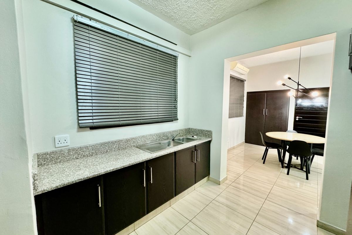 A functional kitchen area featuring dark cabinetry, granite countertops, and a double sink, flowing into an adjacent dining space with tiled flooring throughout.