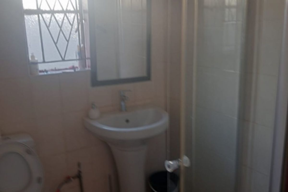 A functional bathroom featuring a pedestal basin, a glass-enclosed shower, and a toilet, with natural light provided by a window with security bars.
