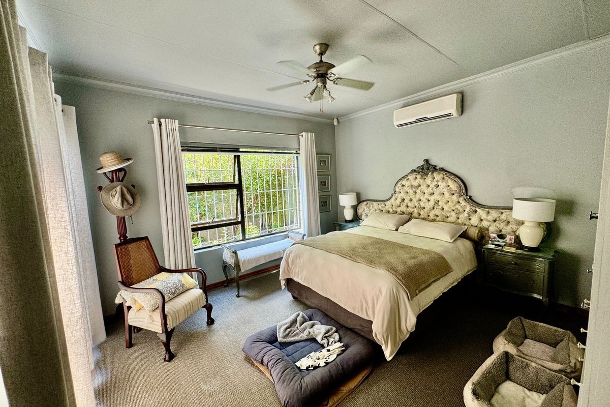 A spacious bedroom featuring a ceiling fan, wall-mounted air conditioning unit, and large window with security bars, providing a comfortable and functional living space.