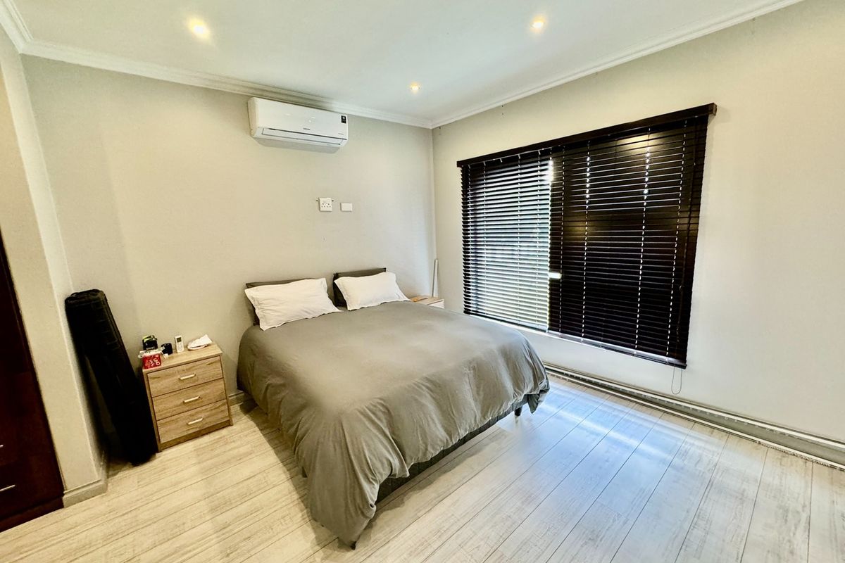A well-maintained bedroom featuring light-toned wood-look flooring, recessed ceiling lighting, a wall-mounted air conditioning unit, and dark timber venetian blinds.