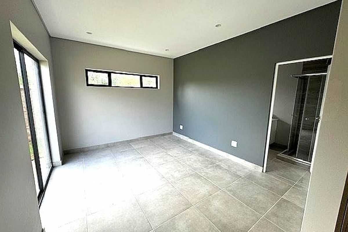 A modern, empty bedroom featuring neutral tiled flooring, recessed ceiling lighting, and an en-suite bathroom with a glass-enclosed shower.