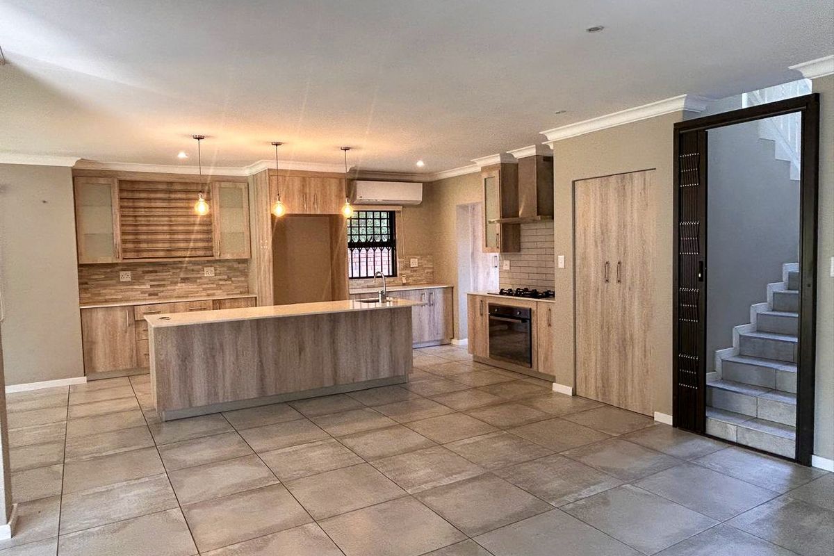 Modern open-plan kitchen featuring wood-finish cabinetry, a central island, built-in oven and hob, and tiled flooring, adjacent to a staircase.
