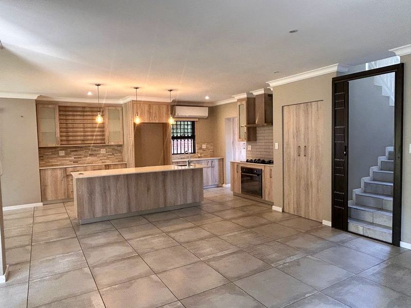 Modern open-plan kitchen featuring wood-finish cabinetry, a central island, built-in oven and hob, and tiled flooring, adjacent to a staircase.