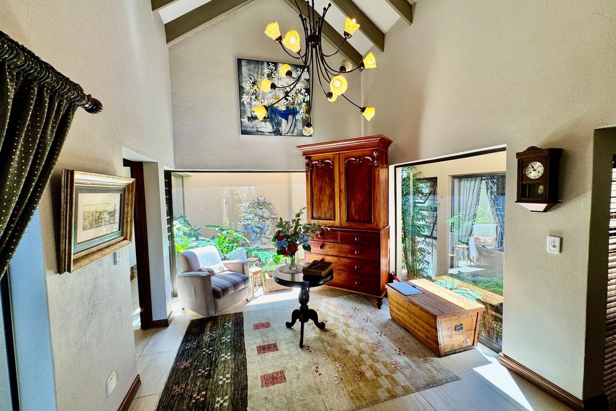 A bright living area featuring a high vaulted ceiling with exposed beams, large windows overlooking a garden, and a decorative chandelier.