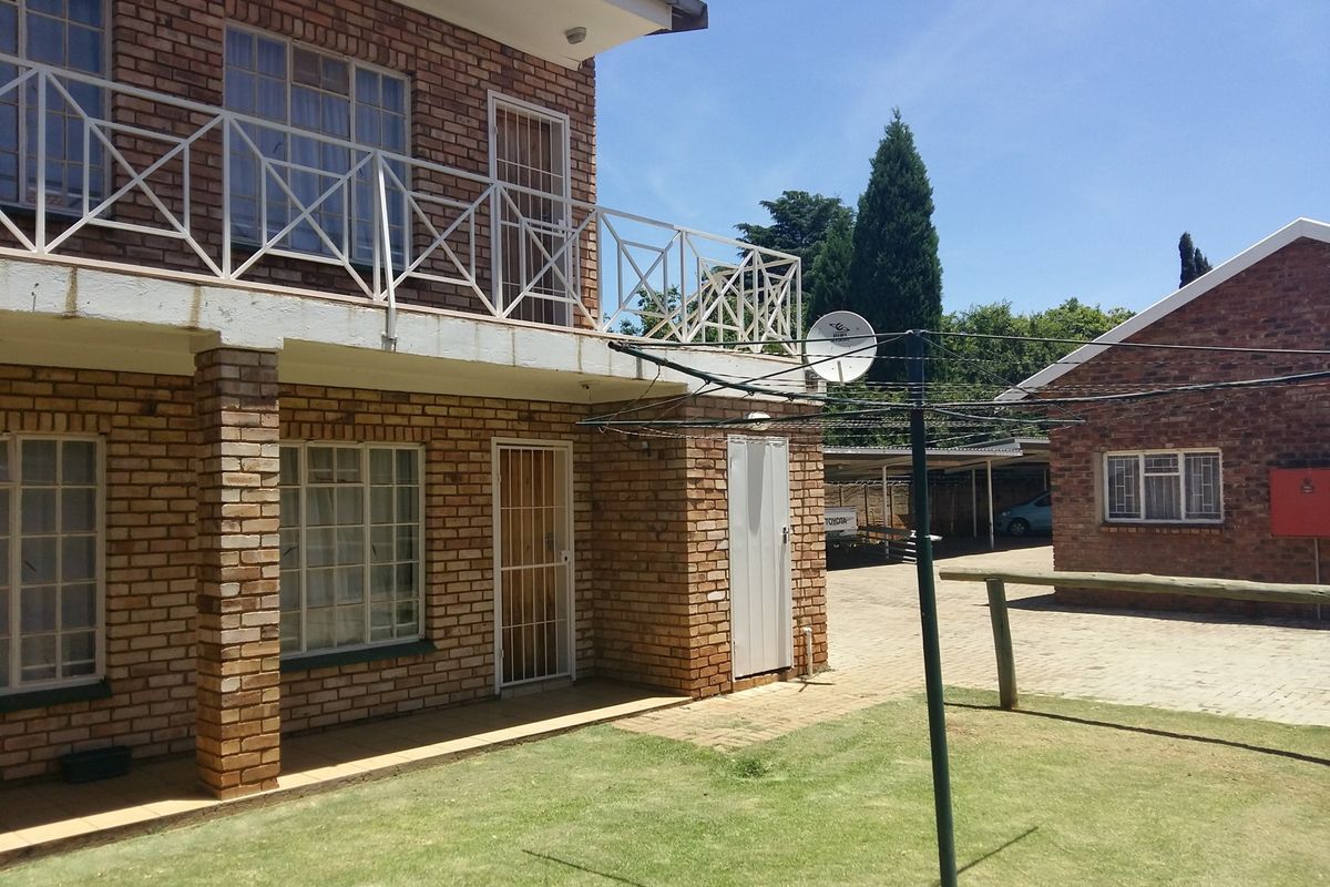 Rear view of a double-story face brick house featuring a balcony with metal railings, a paved driveway, and a small lawn area.