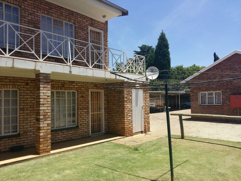 Rear view of a double-story face brick house featuring a balcony with metal railings, a paved driveway, and a small lawn area.