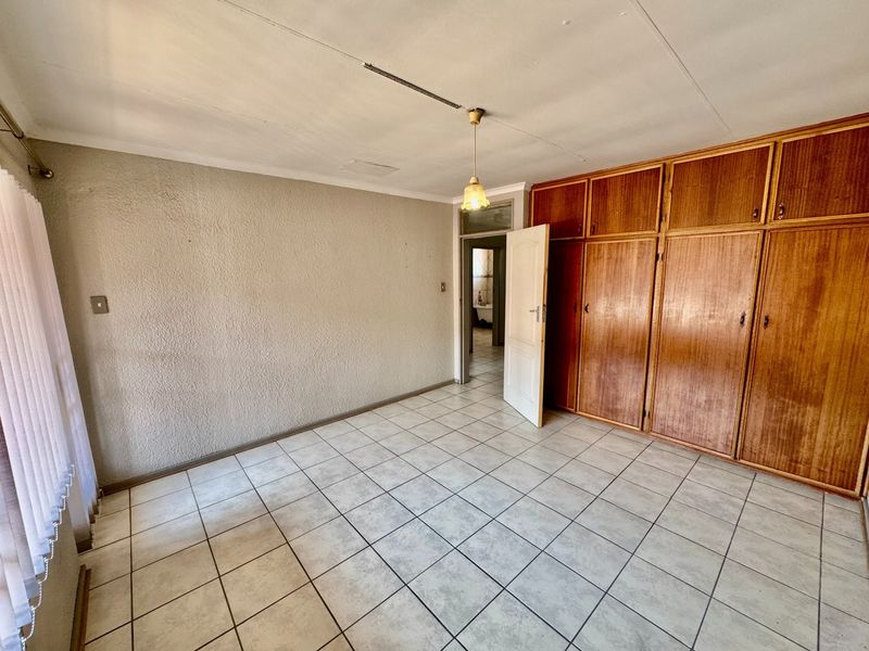 A spacious, empty bedroom featuring tiled flooring, built-in wooden cupboards, and textured walls, offering a blank canvas for renovation.