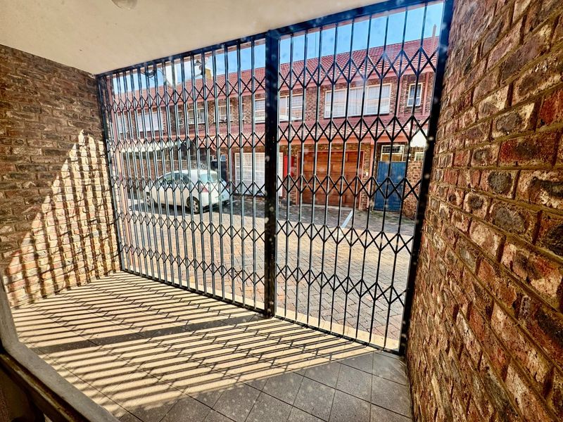 A covered patio area featuring exposed brick walls, tiled flooring, and a secure metal gate looking out towards the townhouse complex driveway.