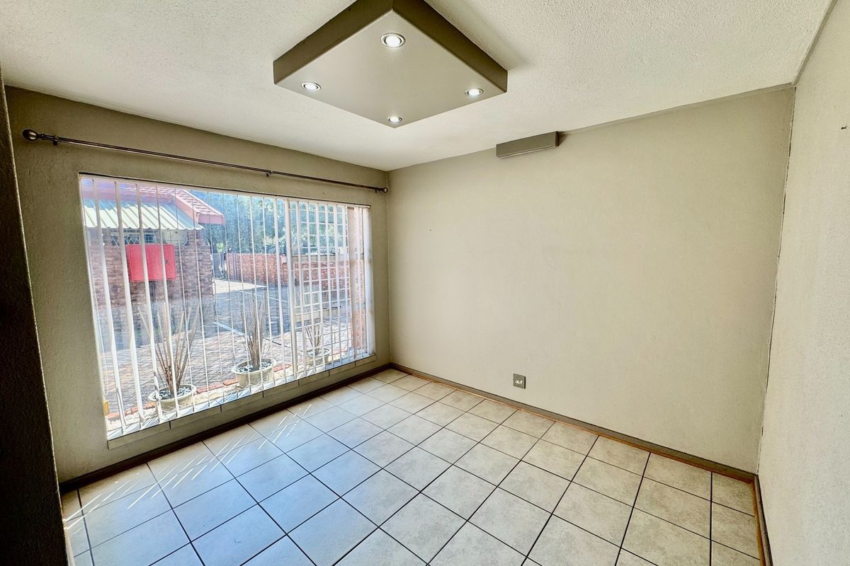 A bright, empty bedroom featuring neutral tiled flooring, a modern recessed ceiling light, and a large window fitted with security bars.