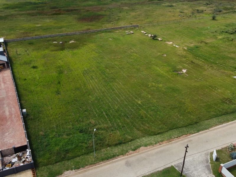 Aerial view of a vacant residential plot featuring a grassed surface, boundary wall, and adjacent paved driveway access.