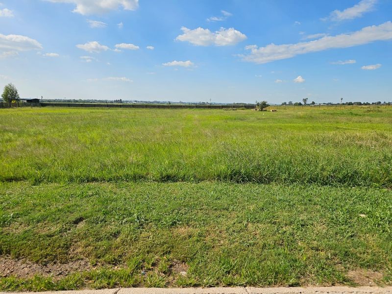 A wide, open residential vacant land plot featuring natural grass coverage and perimeter fencing under a clear blue sky.