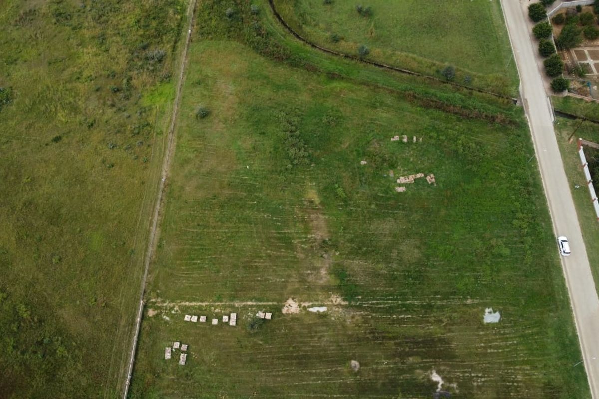 An aerial view of a vacant residential plot featuring open grassland and established boundary lines, situated adjacent to a paved road.