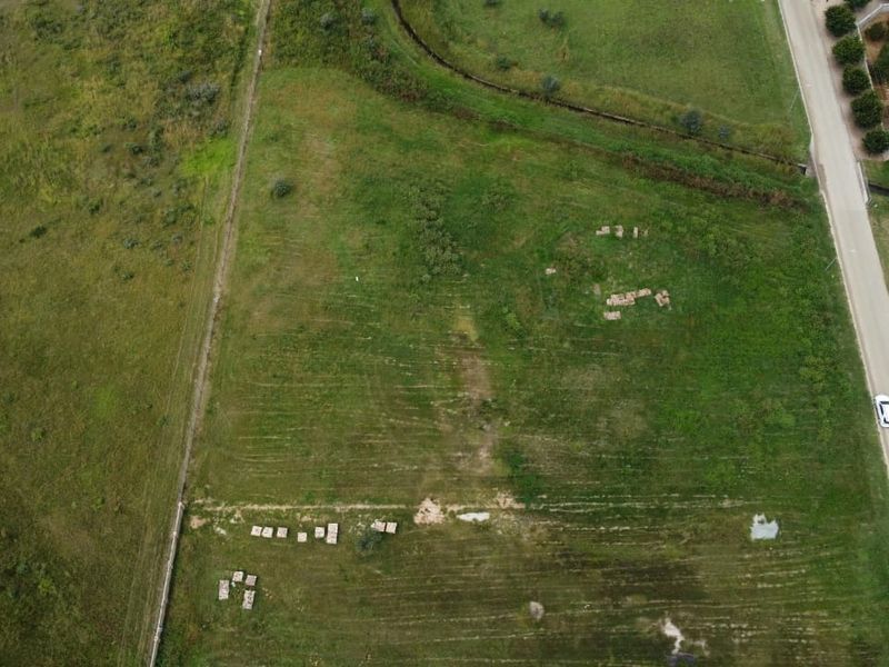 An aerial view of a vacant residential plot featuring open grassland and established boundary lines, situated adjacent to a paved road.