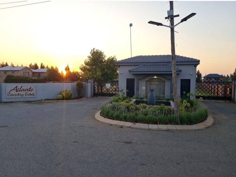 The entrance to Ailanto Country Estate featuring a secure gated access point, perimeter walling, and a landscaped garden with a central water feature.