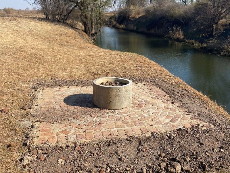 A riverside garden area featuring a circular concrete fire pit set on a cobblestone paved base, overlooking a natural stream.