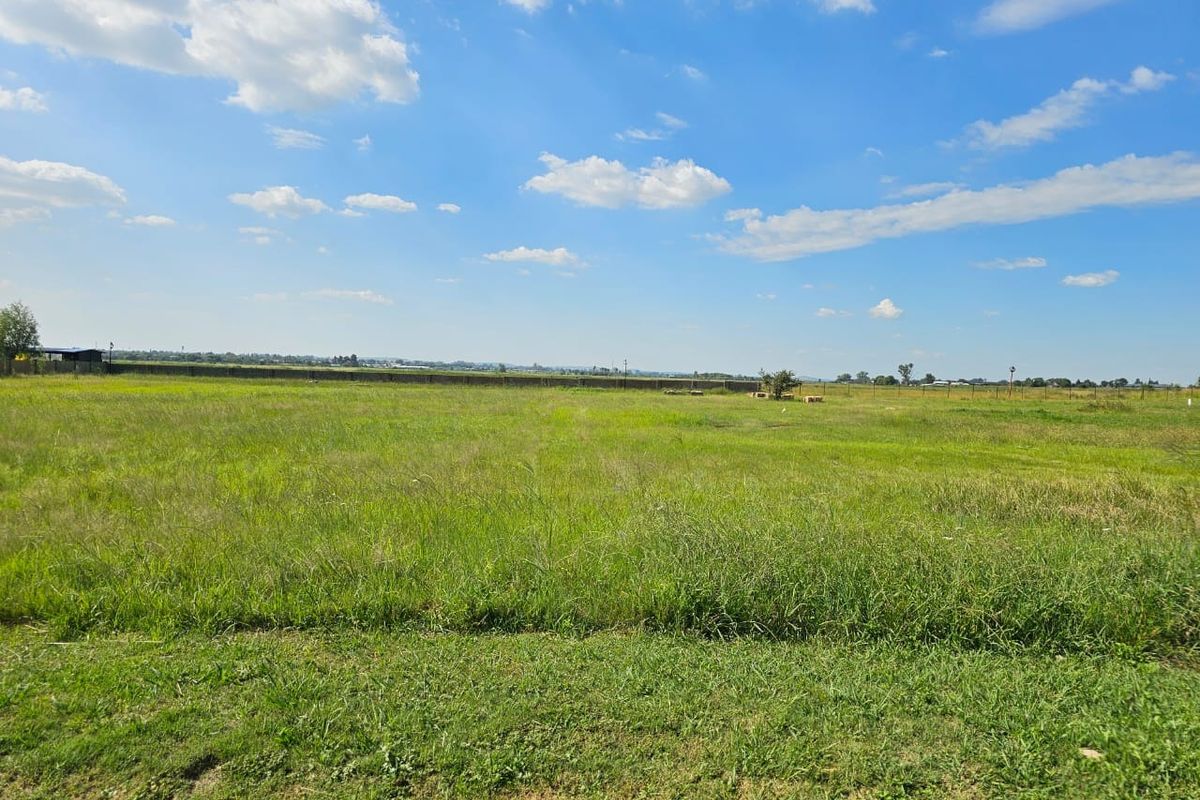 A wide, open vacant residential plot featuring level grassland and perimeter fencing under a clear blue sky.