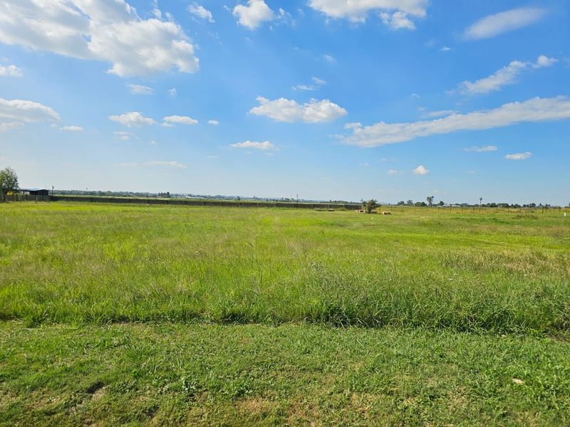 A wide, open vacant residential plot featuring level grassland and perimeter fencing under a clear blue sky.