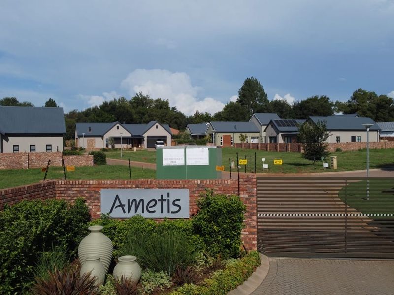 Entrance view of Ametis residential estate featuring a brick boundary wall, metal security gate, and well-maintained landscaped garden.