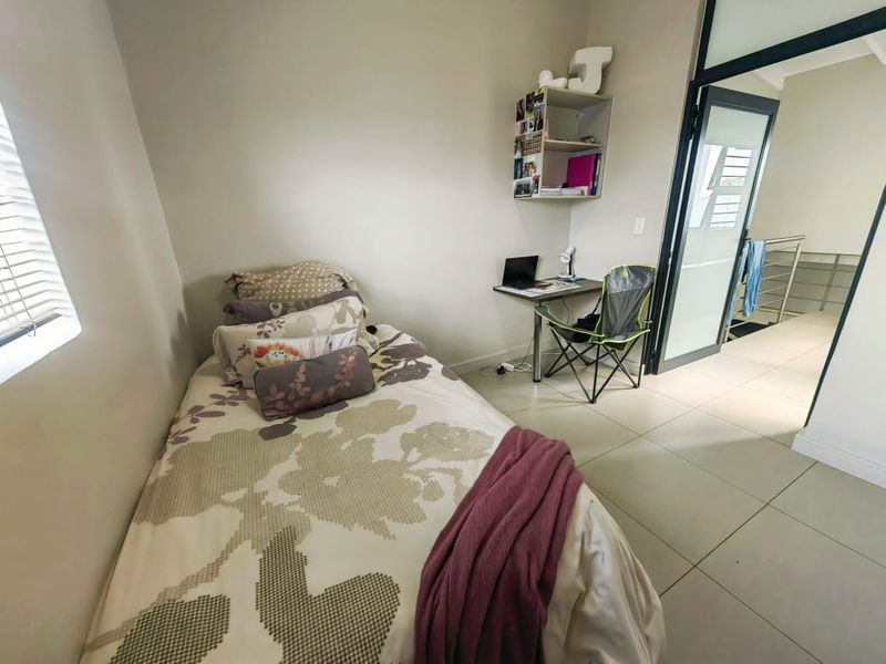 A compact bedroom featuring neutral tiled flooring, venetian blinds, and a modern glass-paneled door leading to an adjacent hallway.