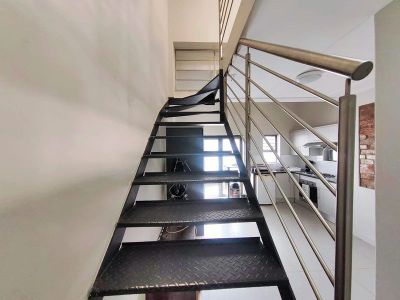 Open plan apartment interior featuring a metal staircase with stainless steel balustrade, leading to a kitchen area with built-in appliances and exposed brick accents.