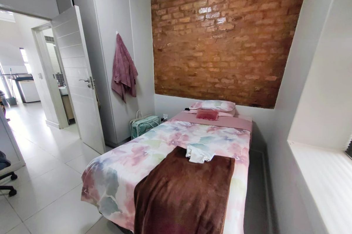 A compact bedroom featuring an exposed brick accent wall, tiled flooring, and a built-in cupboard, with an adjacent view into the hallway.