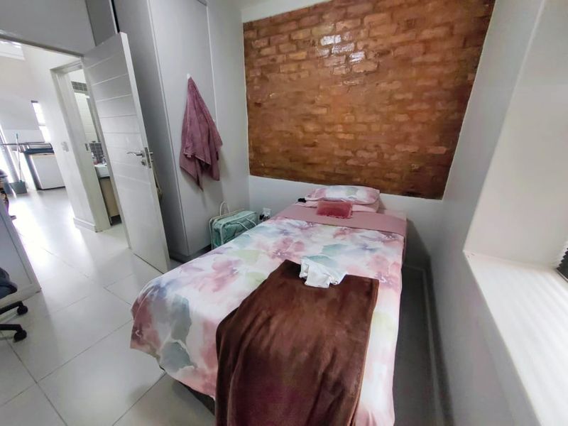 A compact bedroom featuring an exposed brick accent wall, tiled flooring, and a built-in cupboard, with an adjacent view into the hallway.