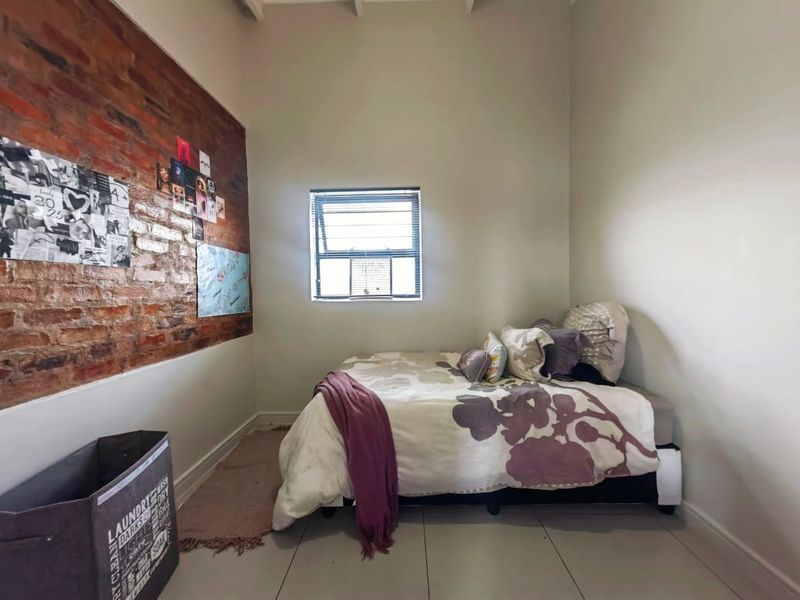 A bedroom featuring an exposed brick feature wall, neutral tiled flooring, and a functional awning window providing natural light.