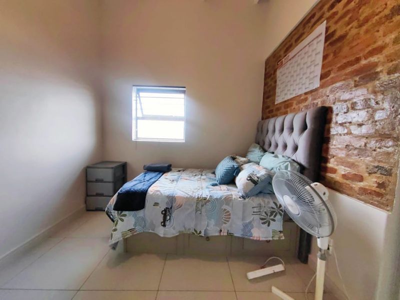 A bedroom featuring a rustic exposed brick feature wall, neutral tiled flooring, and a window providing natural light.