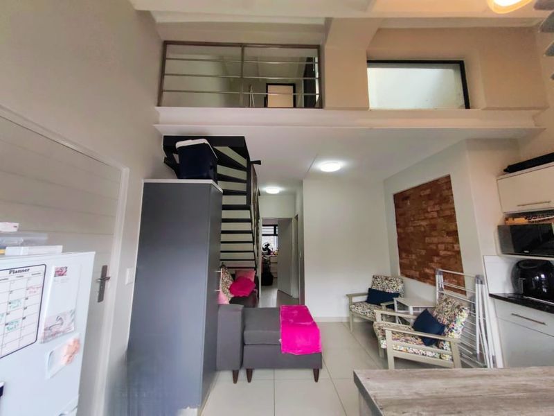 Open-plan living area featuring a mezzanine level, exposed brick accent wall, tiled floors, and a modern staircase leading to the upper floor.