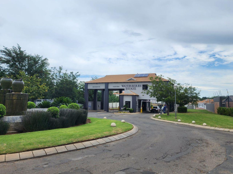 The entrance to Waterberry Estate featuring a secure gatehouse, landscaped gardens, a decorative water feature, and paved access roads.