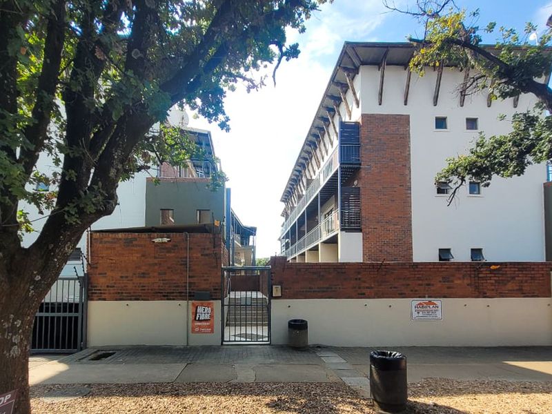 Street-level view of the apartment complex entrance, featuring a brick boundary wall, secure metal gate, and multi-story building facade with balconies.