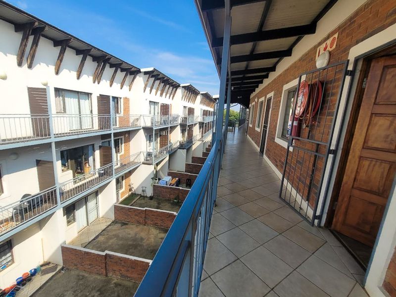 An exterior view of an apartment complex walkway featuring tiled flooring, metal balustrades, and exposed brick walls with a mounted fire hose reel.