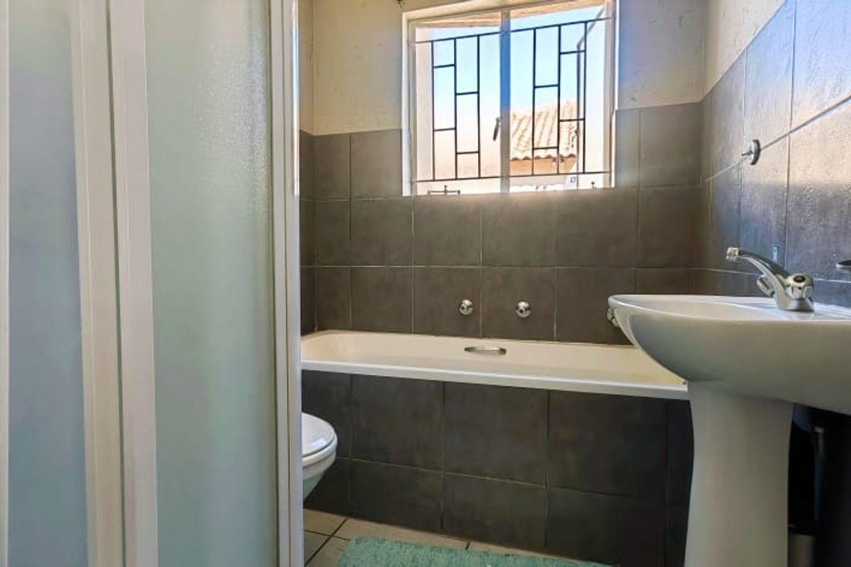 A functional bathroom featuring a bathtub, pedestal basin, and shower enclosure with dark grey wall tiling and a window with security bars.