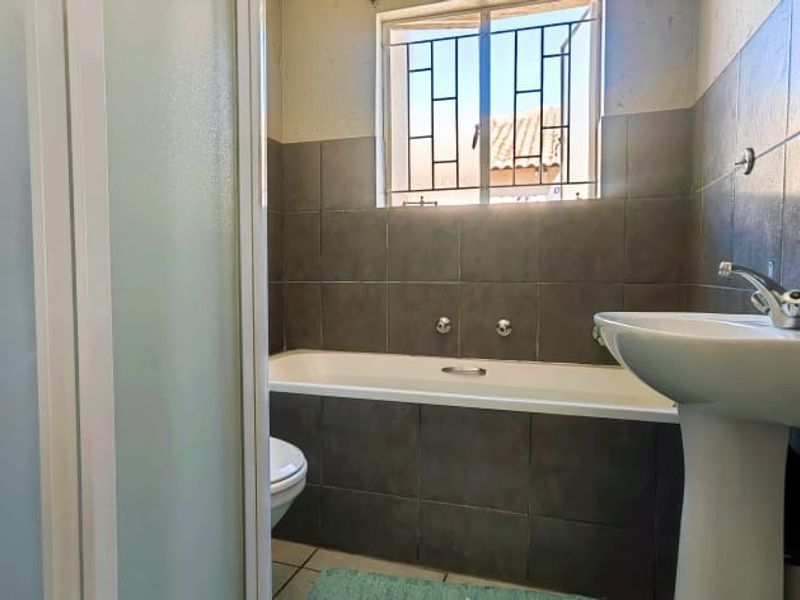 A functional bathroom featuring a bathtub, pedestal basin, and shower enclosure with dark grey wall tiling and a window with security bars.