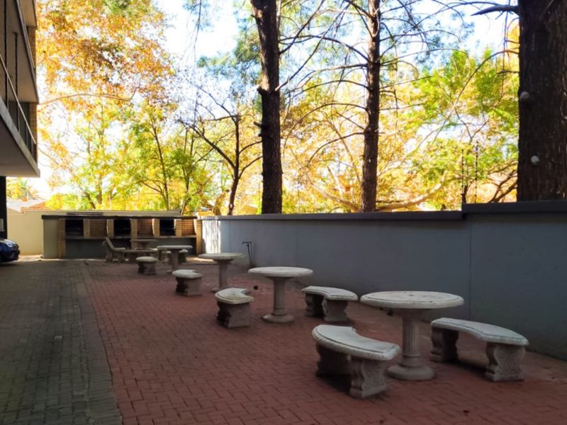 Communal outdoor garden area featuring brick paving, a perimeter wall, and built-in braai facilities set amongst mature trees.