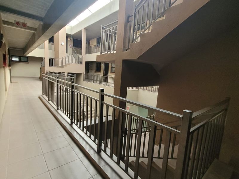 An open-air apartment complex hallway featuring tiled walkways, metal safety balustrades, and a central staircase with overhead skylight lighting.